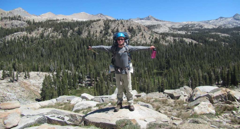 A person wearing safety gear spreads their arms out wide on a rocky overlook. Behind them there is a forest of evergreen trees and a mountain range. 